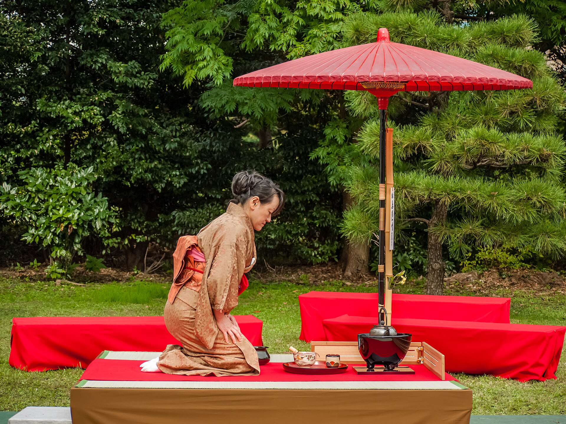 A woman in a brown kimono kneels to prepare tea beneath a red parasol in a garden, with a kettle and lacquer bowls beside her.